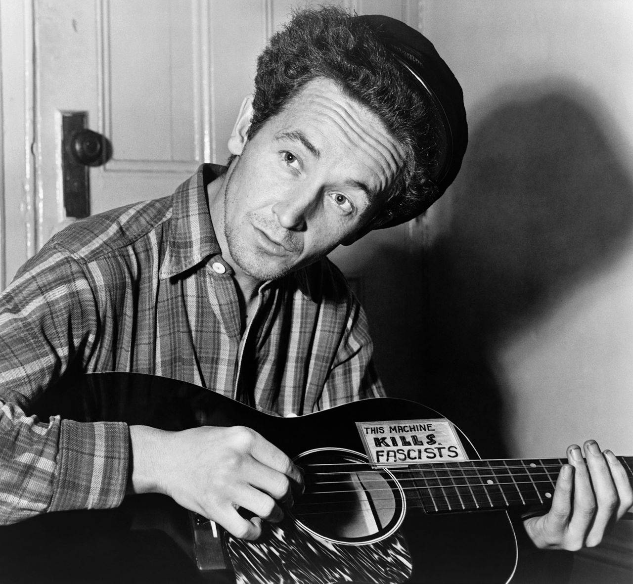 Photograph of Woody Guthrie playing a guitar with a sign that says "This machine kills fascists"