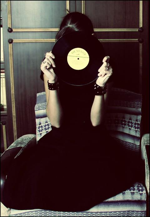Photo of a woman in a black dress holding a vinyl record up to her head, covering her face.