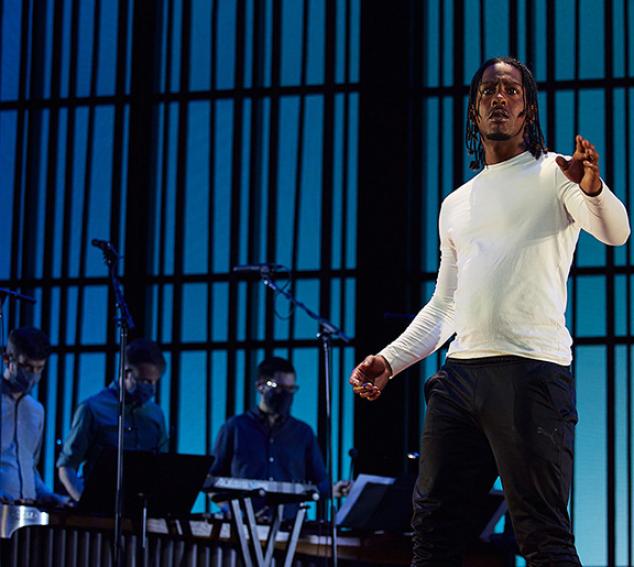 Man in white shirt in front of three musicians on stage with blue pattern background