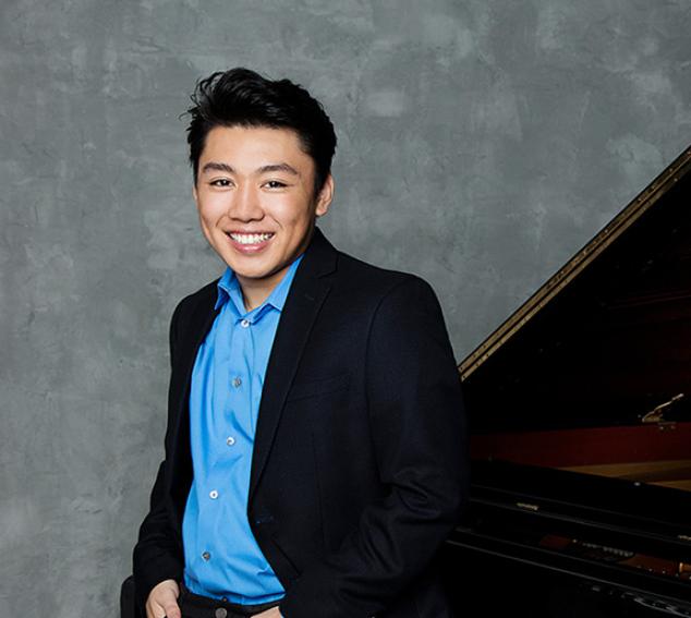 Man in a suit jacket posing in front of a piano and a neutral background