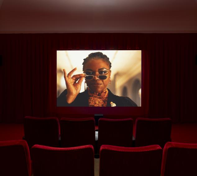 A dark red theater room with red theater-style chairs in the foreground and a screen in the background. The screen portrays a black woman wearing sunglasses looking at the camera.