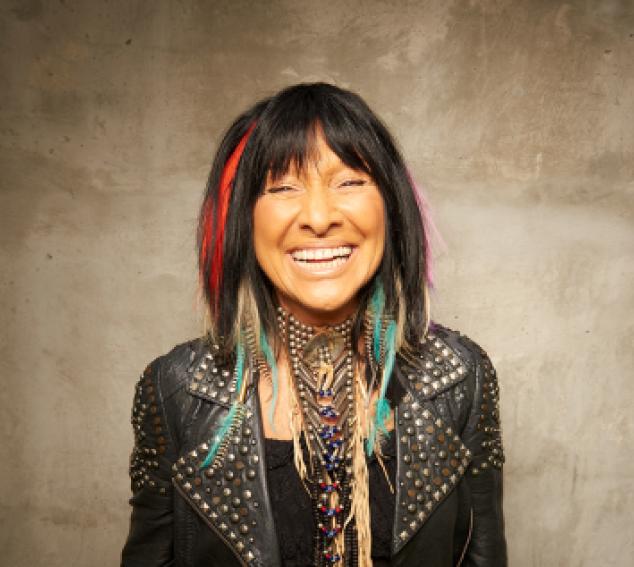 Buffy Sainte-Marie (a smiling woman with dark hair and leather jacket against a tan background)