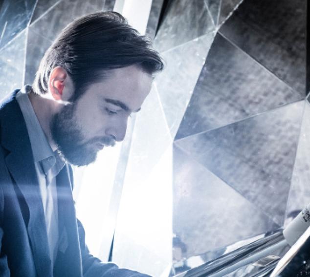 The performer sits at a piano against a gray background