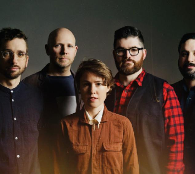 Five performers stand in front of a dark background, looking at the camera