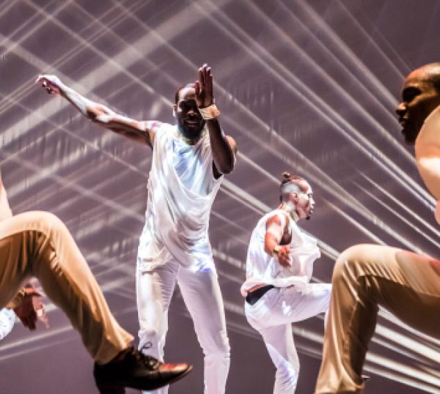 Several dancers perform on a stage with lights in the background